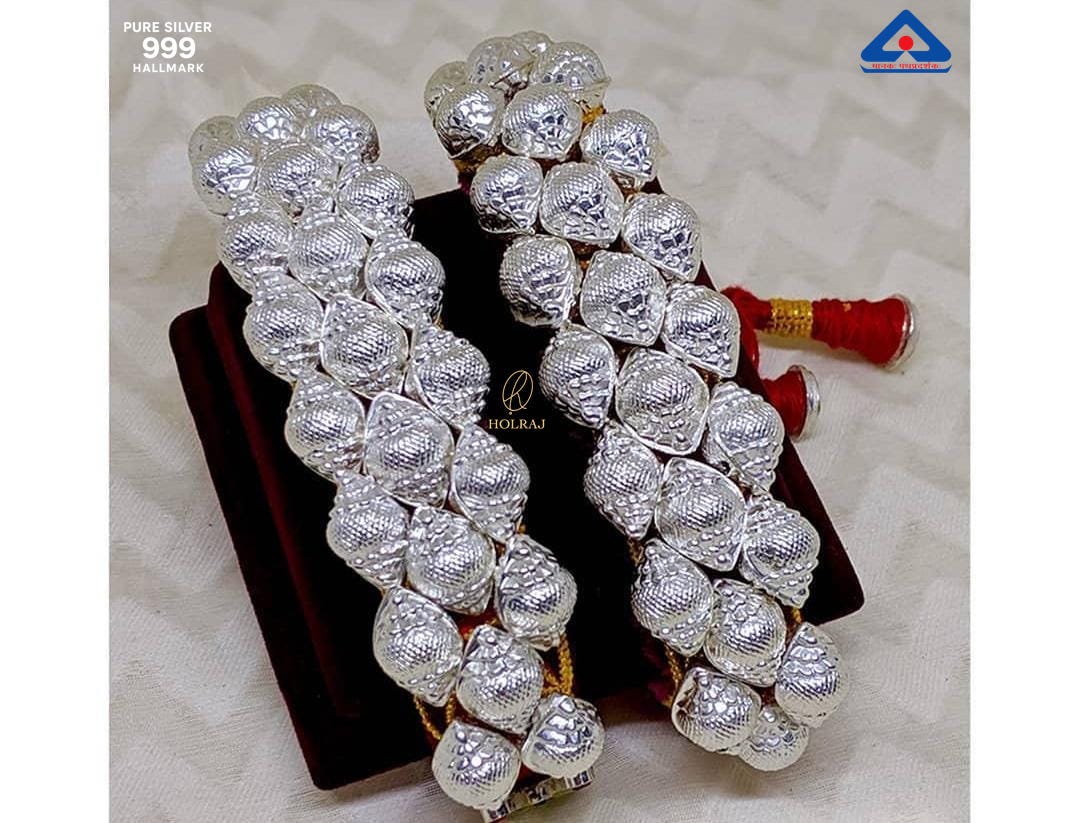 Pair of Shankh Ponchi bracelets in 999 pure silver featuring mirror-polished conch domes on red and gold thread, placed on a soft cream fabric background. By Holraj.
