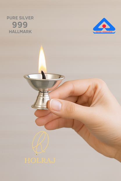 Hand holding a silver 999 hallmark Diwali diya lamp with a flame against a beige background.