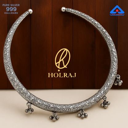 Close-up of a handcrafted 999 silver Hansuli necklace with embossed patterns and ghungroos, placed against a wooden background by Holraj